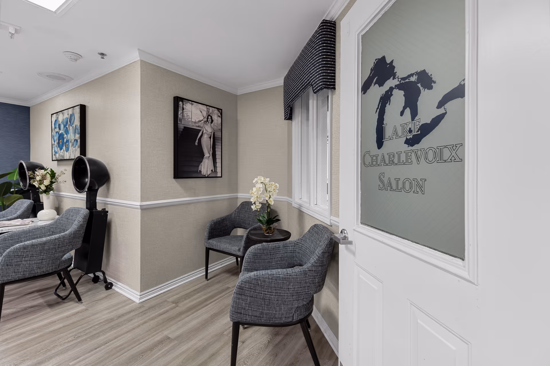 Interior view of a salon waiting area with two gray upholstered chairs and a small round table with a white orchid plant. The door has frosted glass with the text 'Lake Charlevoix Salon'. The room has light-colored walls with white trim, wood flooring, and framed artwork on the walls. Two black hair dryer chairs are visible in the background.