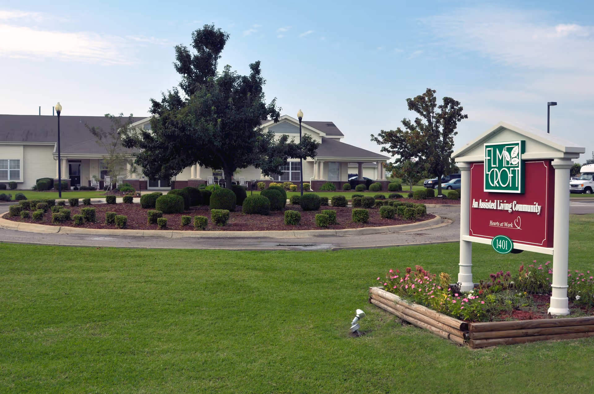 Exterior view of a senior living community with a landscaped roundabout featuring trimmed bushes and a large tree in the center. In the foreground, there is a sign for Elm Croft, an assisted living community, with flowers planted around its base. The building is visible in the background under a partly cloudy sky.