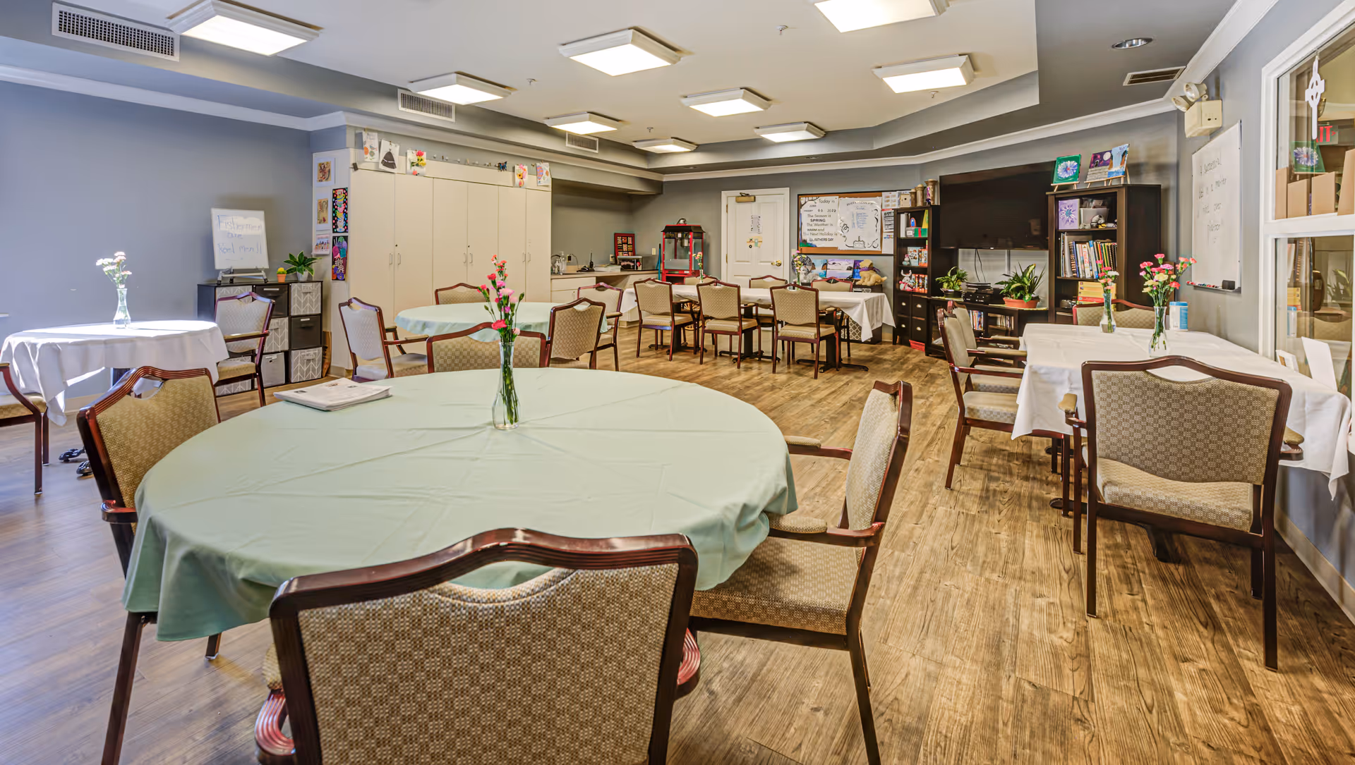 A well-lit community room with multiple round and rectangular tables covered with tablecloths and decorated with small vases of flowers. The room has wooden flooring, a large TV mounted on the wall, bookshelves, and cabinets. There are various decorations and notices on the walls, and the overall setting suggests a comfortable gathering space.