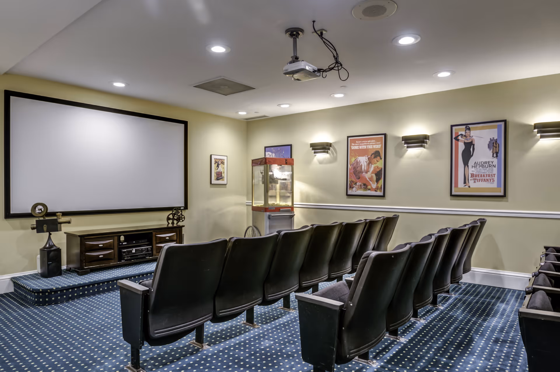 A small movie theater room with two rows of black theater seats facing a large white projection screen. The room has a blue carpet with white dots, beige walls with framed classic movie posters, wall-mounted lights, a popcorn machine in the corner, and a ceiling-mounted projector.