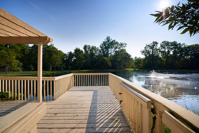 A wooden deck and railing overlooking a calm pond with a small fountain and tree-lined shore under a clear blue sky.