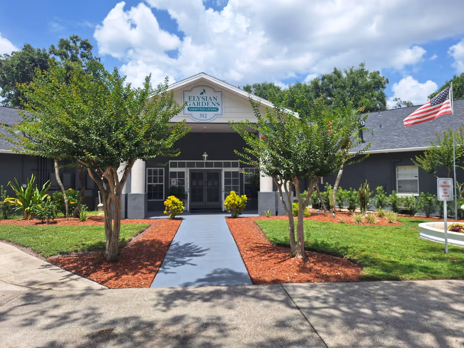 Front exterior view of Elysian Gardens Assisted Living Facility with a pathway leading to the entrance, flanked by small trees and landscaped garden beds. An American flag is visible on the right side under a partly cloudy sky.