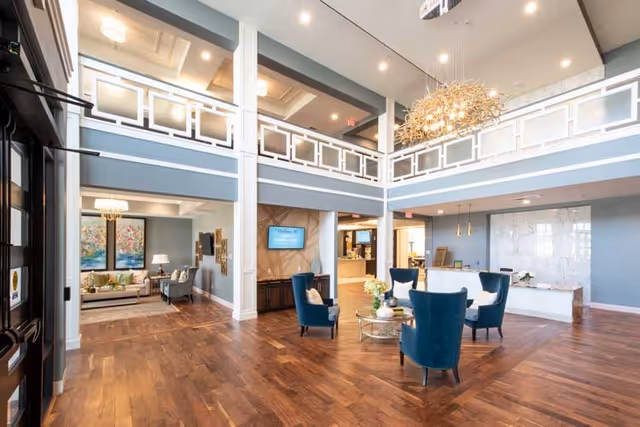 Spacious and well-lit senior living facility lobby with high ceilings, wooden floors, and modern decor. The area features a seating arrangement with blue upholstered chairs around a glass coffee table, a large chandelier, and a reception desk in the background. There is a second-floor balcony with white railings overlooking the lobby, and a cozy sitting area with sofas and artwork is visible to the left.