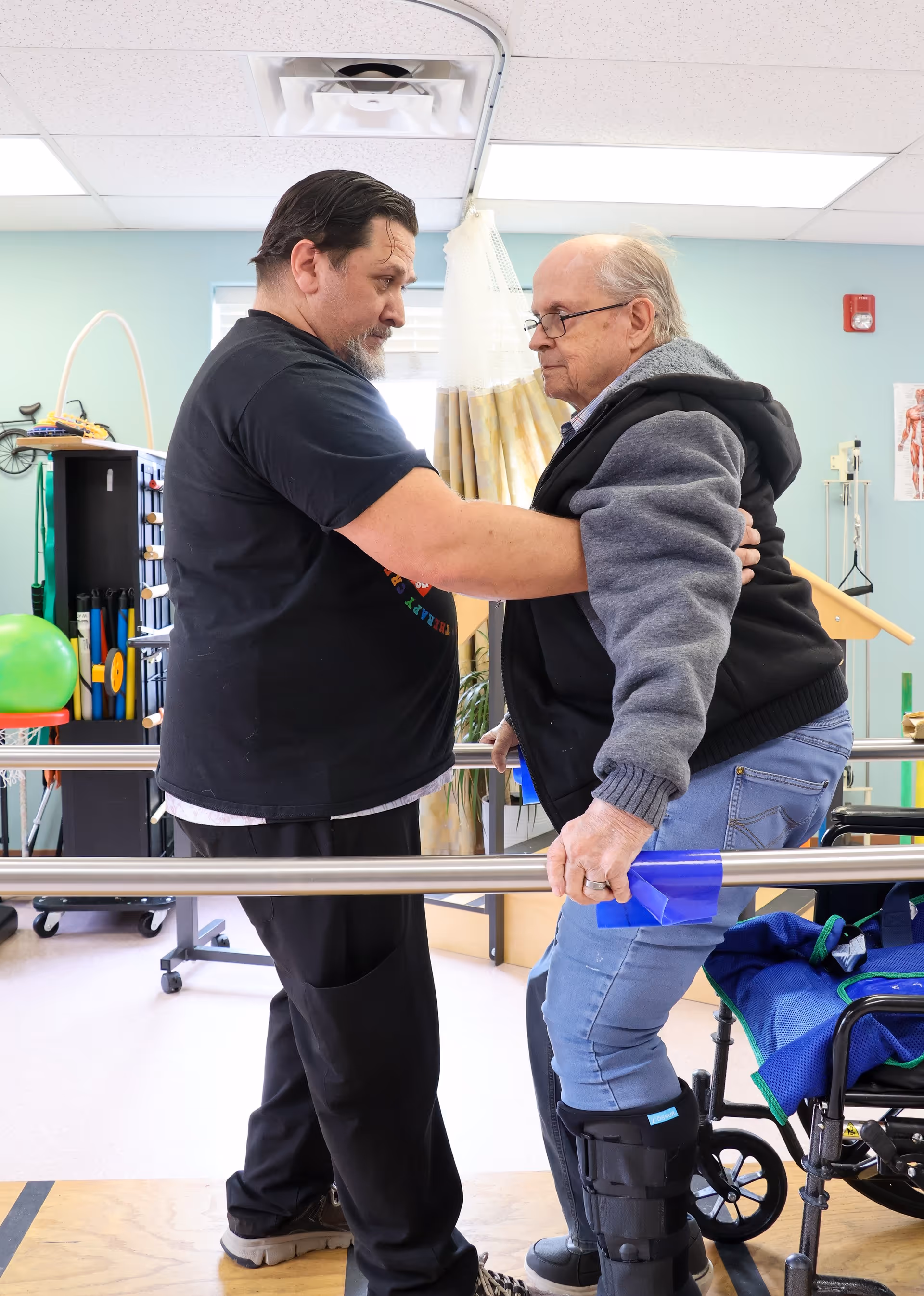 A caregiver assists an elderly man with a leg brace as he practices walking using parallel bars in a rehabilitation or physical therapy room. The elderly man holds onto the bars for support while the caregiver supports him by holding his arm and back. A wheelchair and exercise equipment are visible in the background.