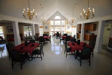 Spacious dining room with red tablecloths, chairs, and chandeliers beneath a large sunlit window.