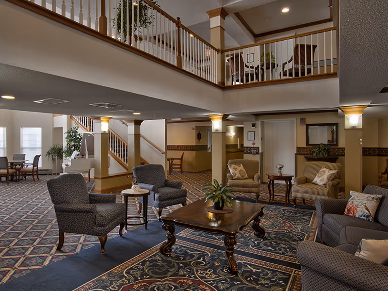 Spacious senior living facility common area with patterned carpet, multiple upholstered chairs and sofas arranged around wooden coffee tables, potted plants, a white piano near large windows, and a staircase leading to an upper balcony with railing.