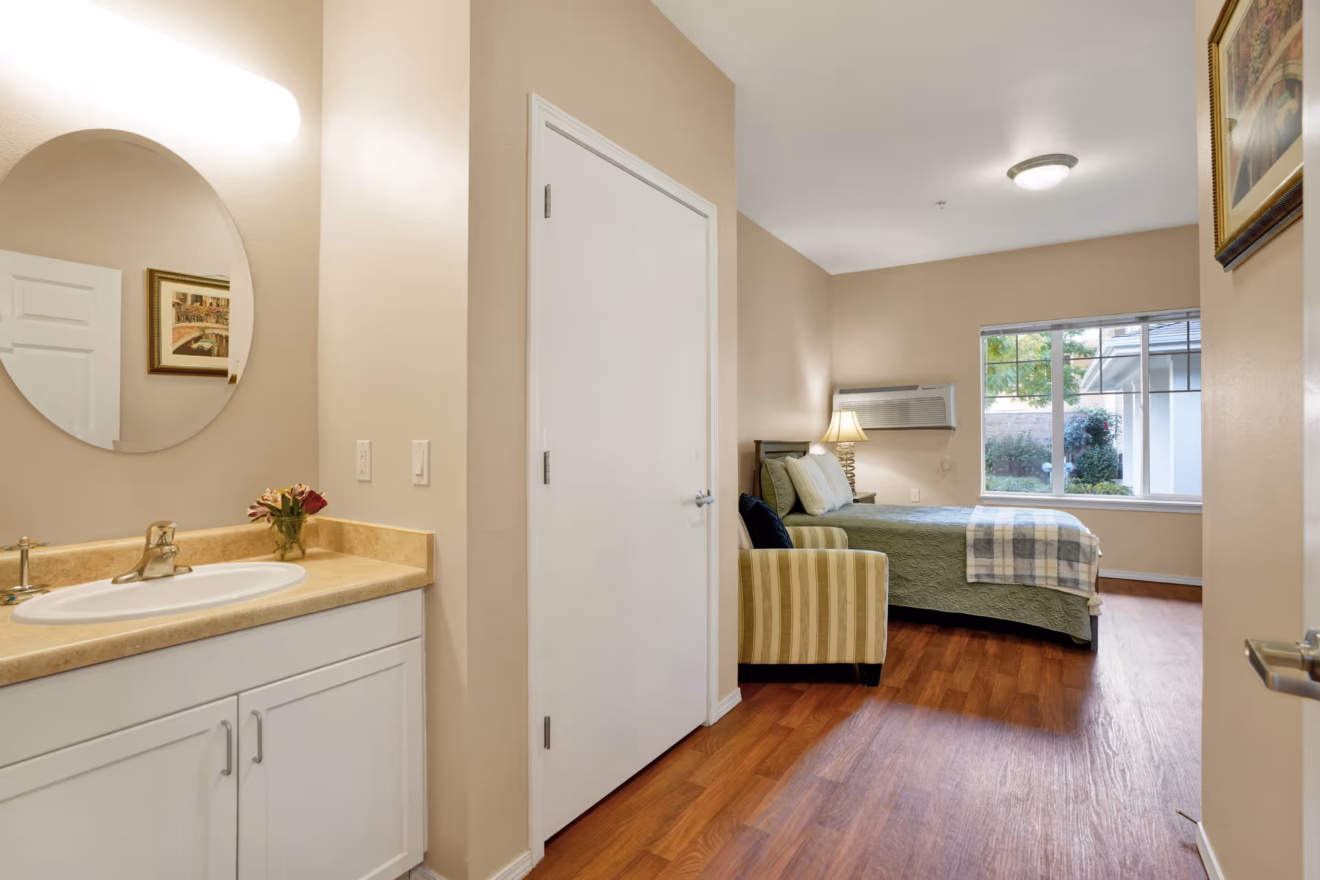 A senior living facility room with a bed covered in a green quilt and a plaid blanket, a striped armchair, a nightstand with a lamp, and a window showing greenery outside. To the left, there is a bathroom sink with a round mirror and a small vase with flowers on the countertop. The room has wooden flooring and beige walls.