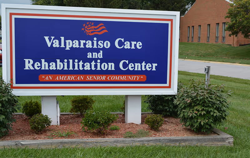 Outdoor sign for Valparaiso Care and Rehabilitation Center with the tagline 'An American Senior Community' displayed on a blue background with red and white borders, surrounded by small bushes and grass, with a brick building and road in the background.