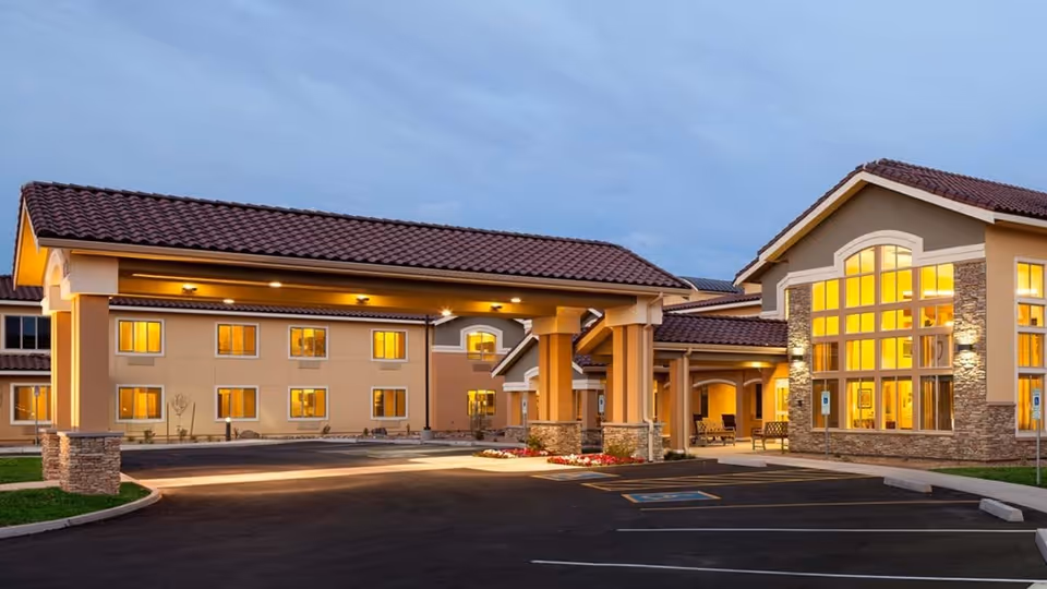 Exterior view of Farmington Station Senior Living facility at dusk, showing a large building with warm yellow lights glowing from windows, a covered entrance with stone pillars, and an empty parking lot with marked handicap spaces.