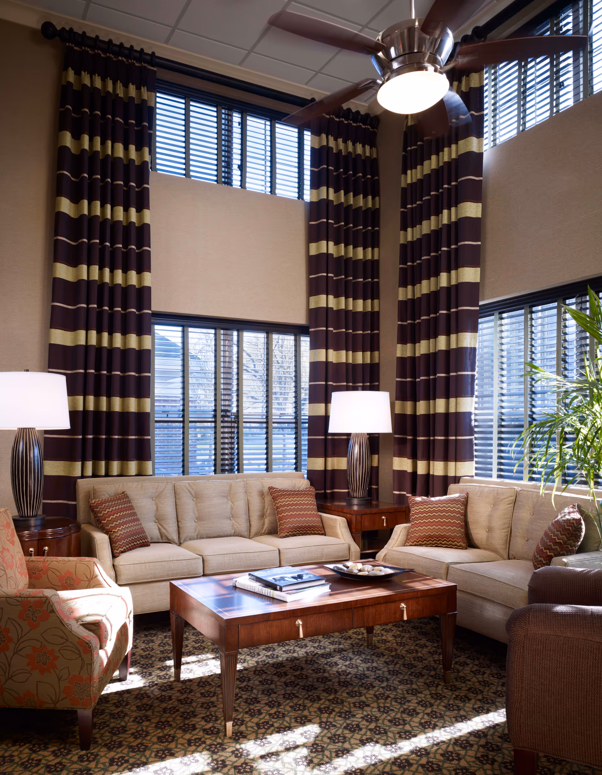 Sunlit communal living room with sofas and armchairs around a wooden coffee table, tall striped curtains, table lamps and a ceiling fan.