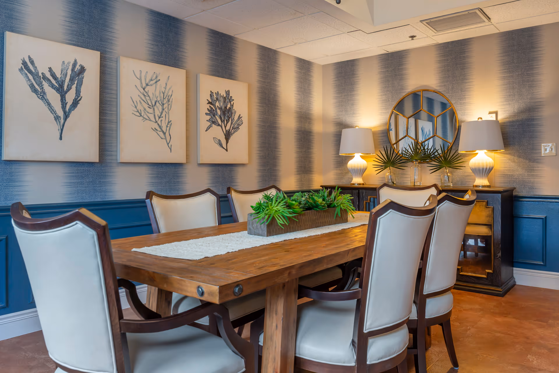 A well-decorated dining room with a wooden table, upholstered chairs, wall art, and lamps on a sideboard.