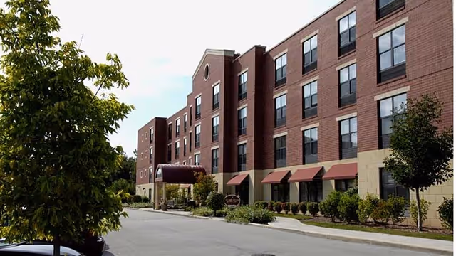 Exterior view of a multi-story brick building with several windows and red awnings over the ground floor windows. There is a covered entrance with a maroon canopy and landscaped greenery including bushes and trees along the sidewalk.