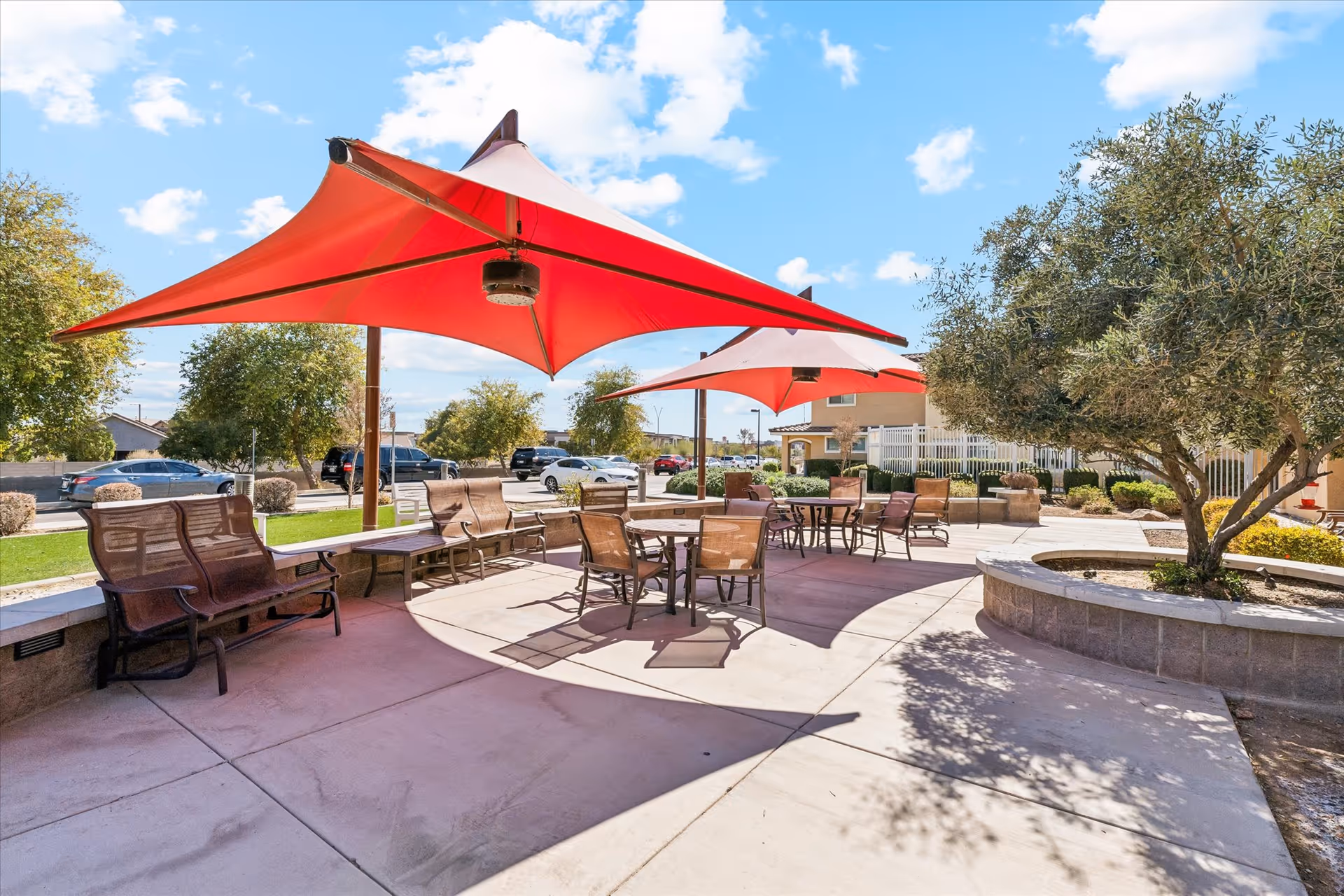 Outdoor patio area with several tables and chairs under large red triangular shade sails. There are benches along the curved stone wall and trees providing additional shade. The sky is blue with some clouds, and cars are parked in the background near a building.