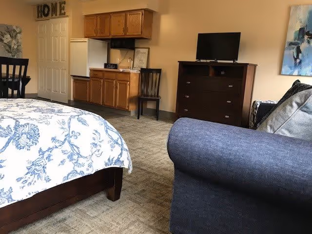 A studio-style senior living room showing a bed with floral bedding, a couch, dresser with TV, small kitchenette and dining area.