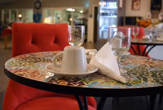 A close-up view of a round table with a colorful mosaic top in a dining area. On the table, there is an upside-down white coffee cup on a saucer, a folded white napkin, and two empty glasses. In the background, there are red cushioned chairs and a refrigerator with beverages inside.