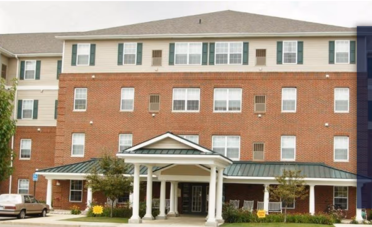 Exterior view of a multi-story senior living facility building with a brick facade on the lower floors and beige siding on the upper floor. The entrance features a covered portico with white columns and a green metal roof. There are several windows with green shutters and a few small trees and shrubs near the entrance. A car is parked on the left side near a handicap parking sign.