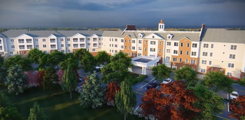 Exterior view of a large senior living facility named Harmony at Kent, featuring a multi-story building with a mix of white and brick facade, surrounded by trees and a parking area in front.
