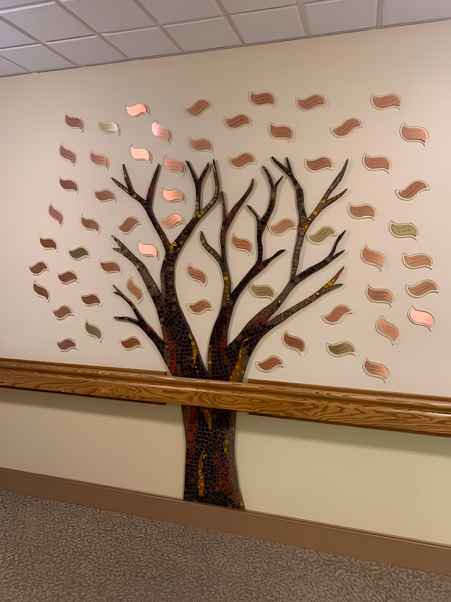 Decorative mosaic tree wall installation with scattered leaf shapes mounted on a hallway wall above a wooden handrail.