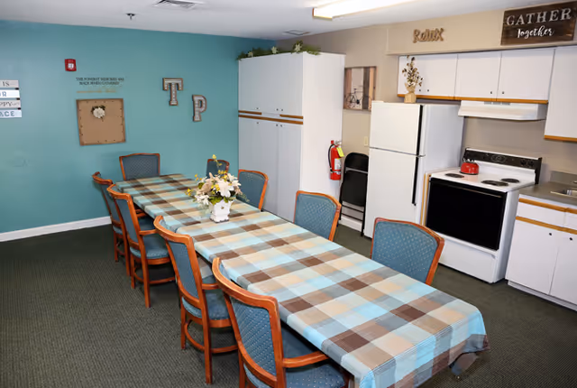 A communal dining room with a long plaid-covered table and chairs beside a small kitchenette and teal accent wall.