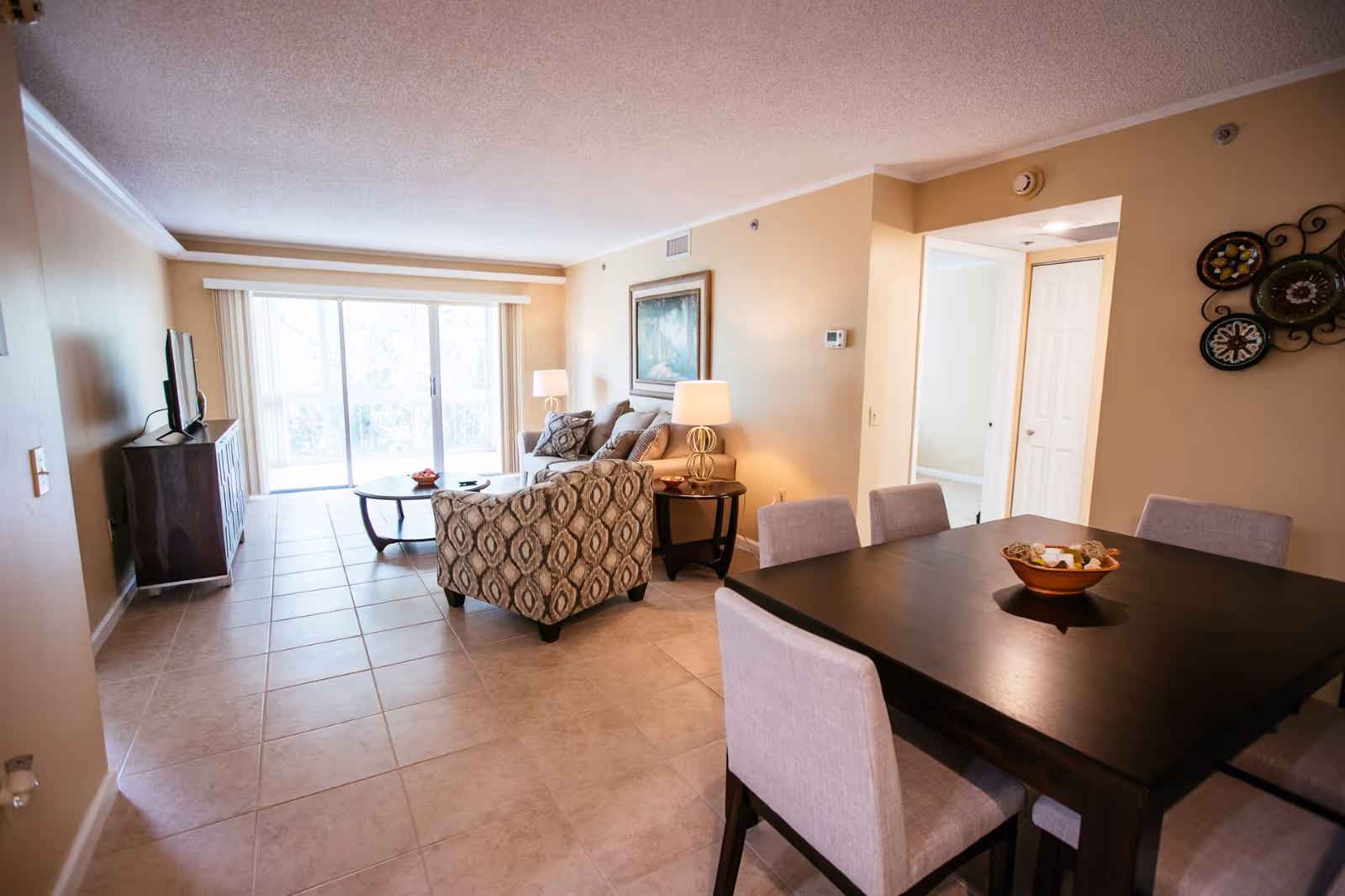 A bright and spacious living and dining area in a senior living facility. The room features a tiled floor, a dark wooden dining table with six light-colored upholstered chairs, a patterned armchair, a beige sofa with cushions, two side tables with lamps, a coffee table, a TV on a stand, and large sliding glass doors leading to a balcony with natural light coming through.