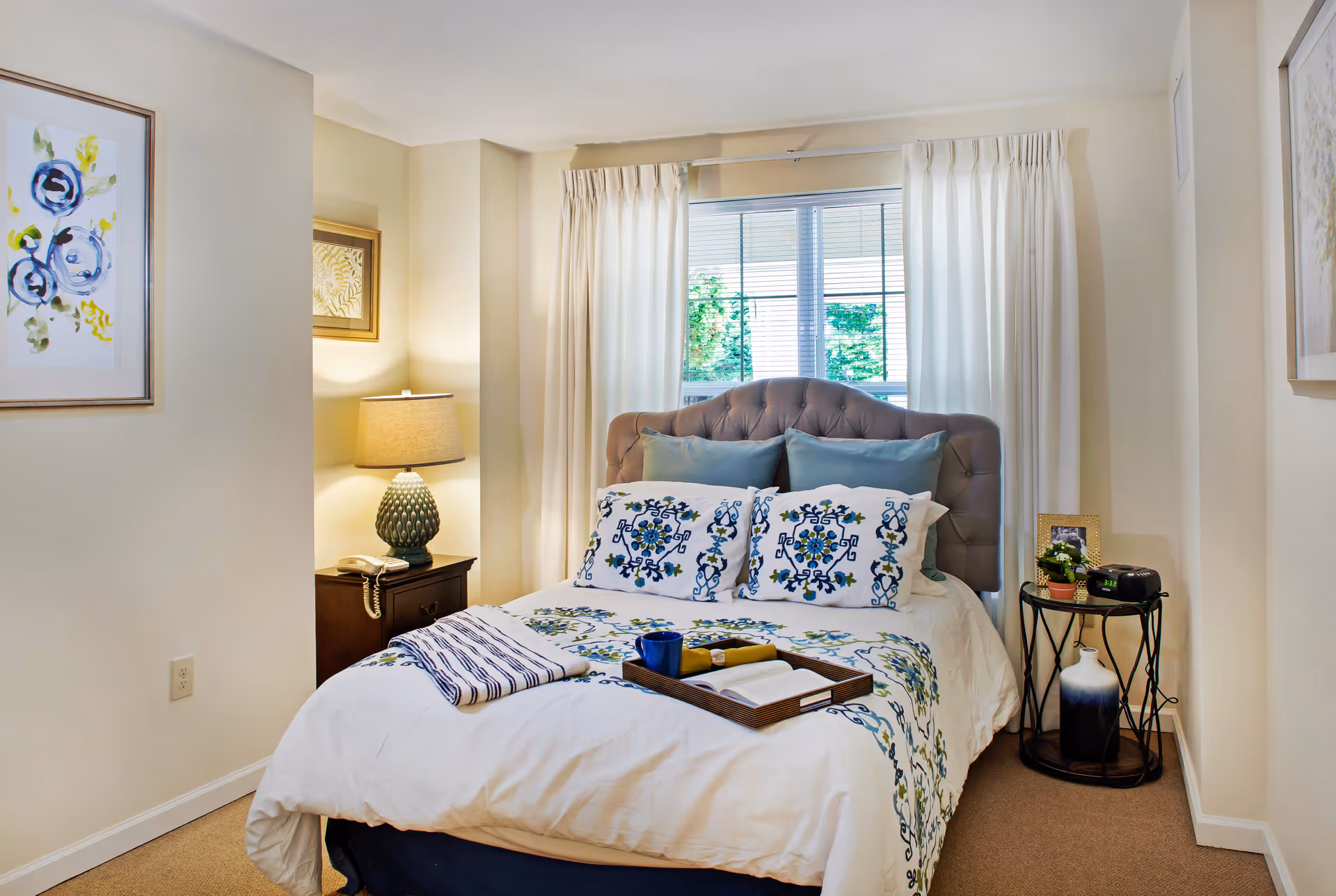 Bright, neatly staged bedroom with a tufted headboard, patterned bedding, bedside tables, and a window with curtains.