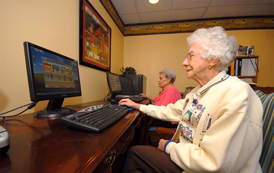 Two elderly women sitting at a wooden desk using desktop computers in a room with yellow walls and framed artwork.