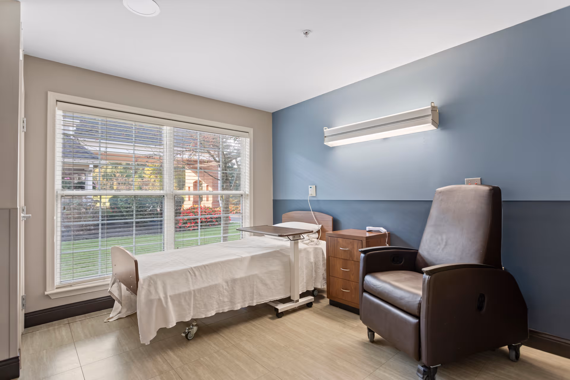 A small, clean room with a single hospital-style bed covered with a white sheet, a wooden bedside table with drawers, a brown leather recliner chair, and a large window with white blinds showing a green lawn and bushes outside. The walls are painted in two tones, beige and blue, and there is a fluorescent light fixture mounted above the bed.