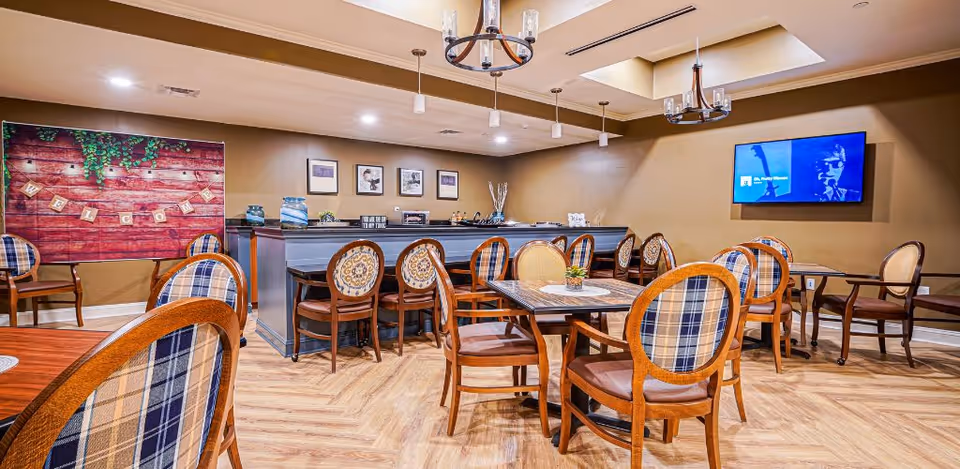 A cozy dining area with several wooden tables and chairs featuring patterned upholstery. The room has warm beige walls, a wooden floor, and two chandeliers hanging from the ceiling. A flat-screen TV is mounted on the wall, and a decorative welcome sign is displayed on the left side. There are framed pictures on the wall behind a counter or bar area.