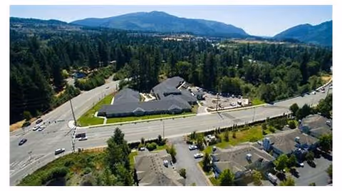 Aerial view of a residential area with a main road running through the center. The image shows multiple buildings surrounded by trees and greenery, with hills and mountains in the background under a clear sky.