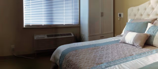 A bedroom with a neatly made bed featuring a tufted headboard, multiple pillows, and a quilted bedspread in shades of blue and beige. Next to the bed is a white wardrobe with drawers, and beneath a window with closed blinds is an air conditioning unit.