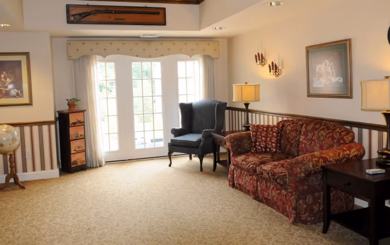 A cozy living room area with a patterned red sofa, a dark blue armchair, two side tables with lamps, a globe on a stand, framed artwork on the walls, and large glass doors letting in natural light.