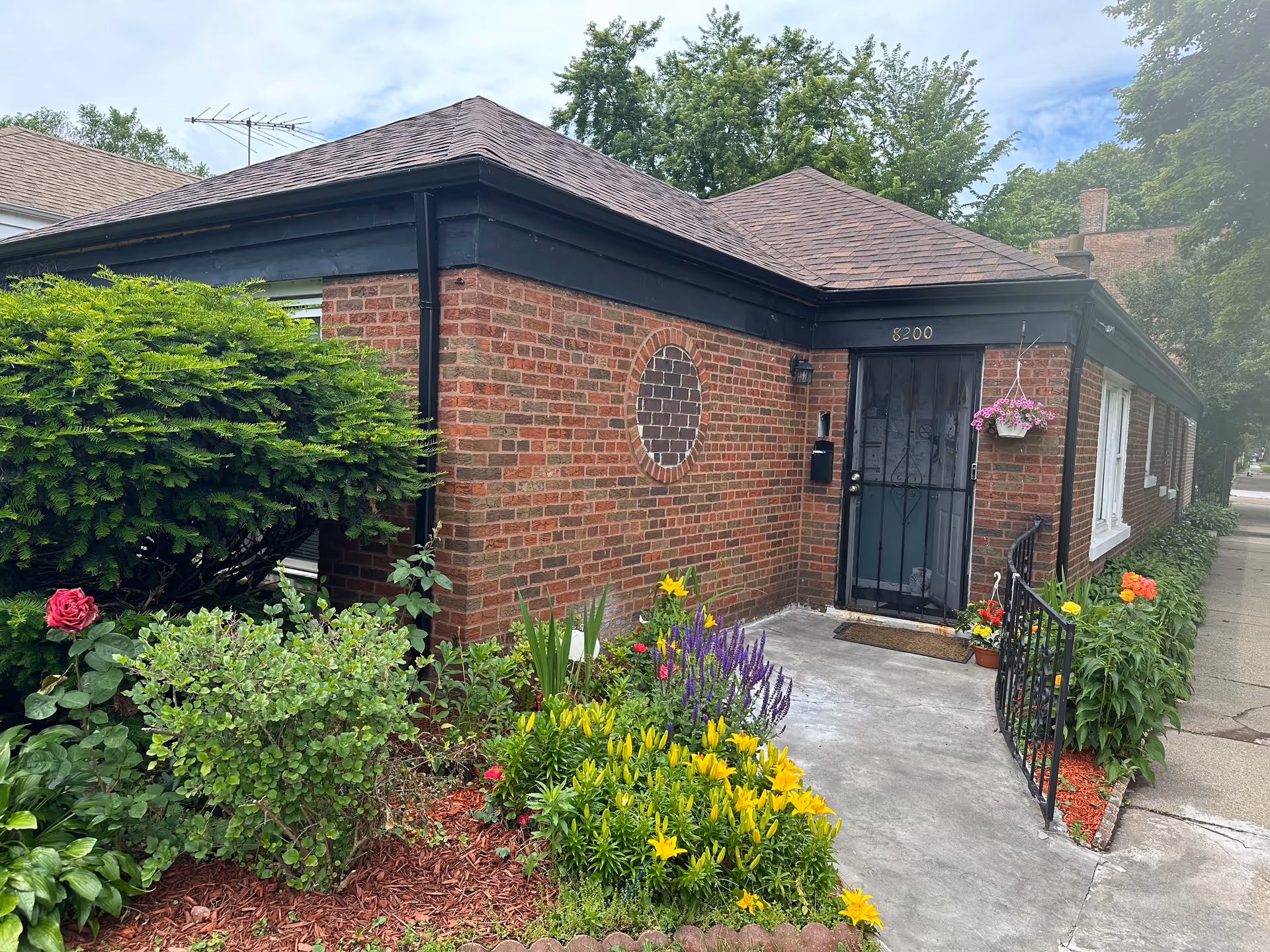 Exterior view of a single-story brick building with a black door and the number 8200 above it. The entrance is surrounded by a small garden with colorful flowers and green shrubs. There is a concrete pathway leading to the door, and a black metal railing on one side. Trees and a partly cloudy sky are visible in the background.