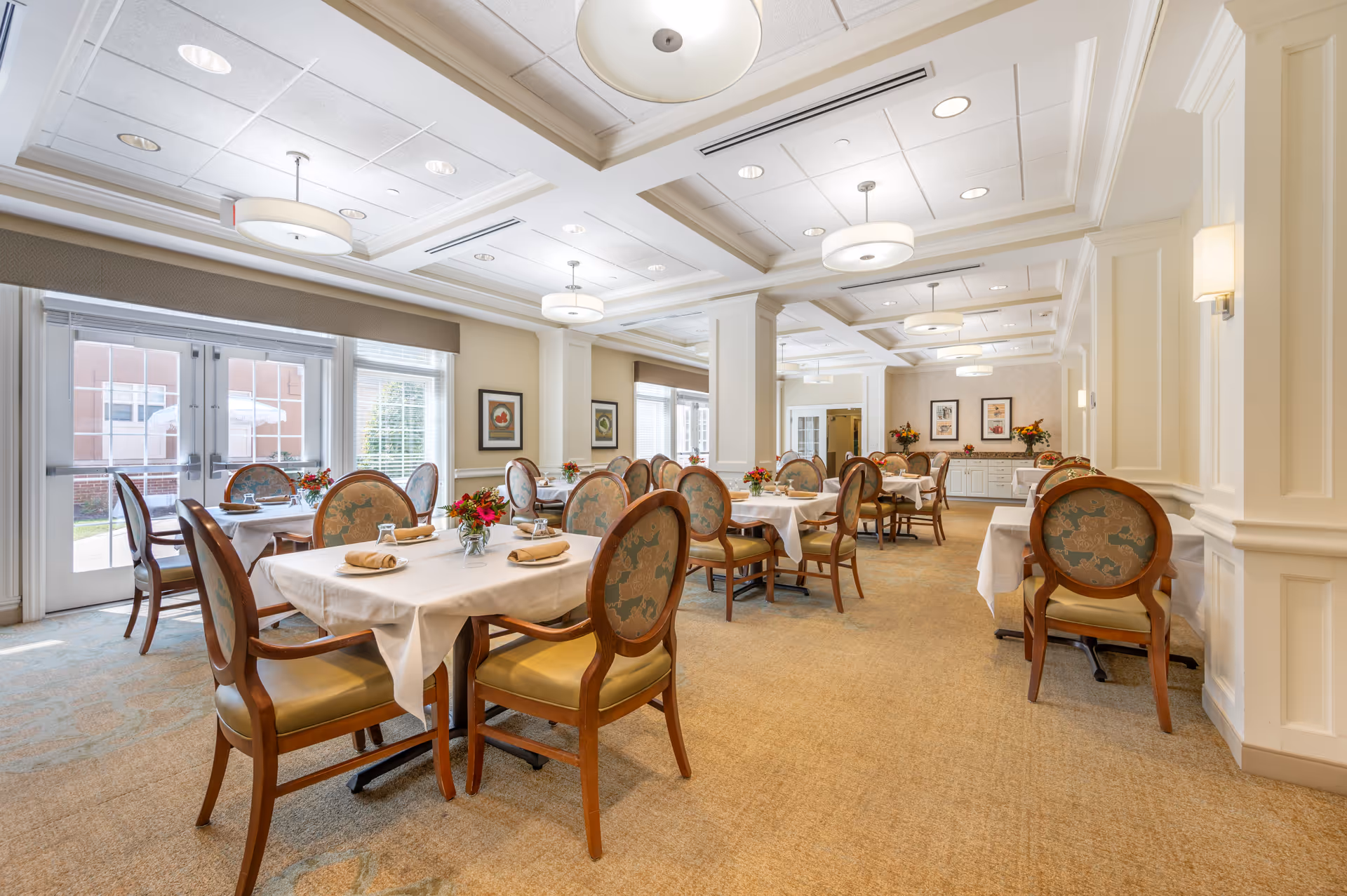 A bright and elegant dining room with multiple tables covered in white tablecloths, each set with napkins, glasses, and small floral centerpieces. The room features cushioned wooden chairs with patterned upholstery, large windows allowing natural light, and ceiling lights with round fixtures. The walls are decorated with framed artwork and the overall ambiance is warm and inviting.