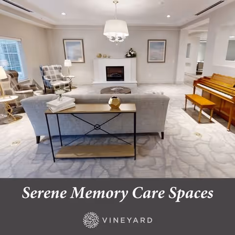 A serene memory care living room with a gray sofa facing a white fireplace. The room features two armchairs with plaid upholstery, a wooden piano with a bench, a side table with a lamp, and framed artwork on the walls. The carpet has a subtle floral pattern, and the space is softly lit with a ceiling light fixture.