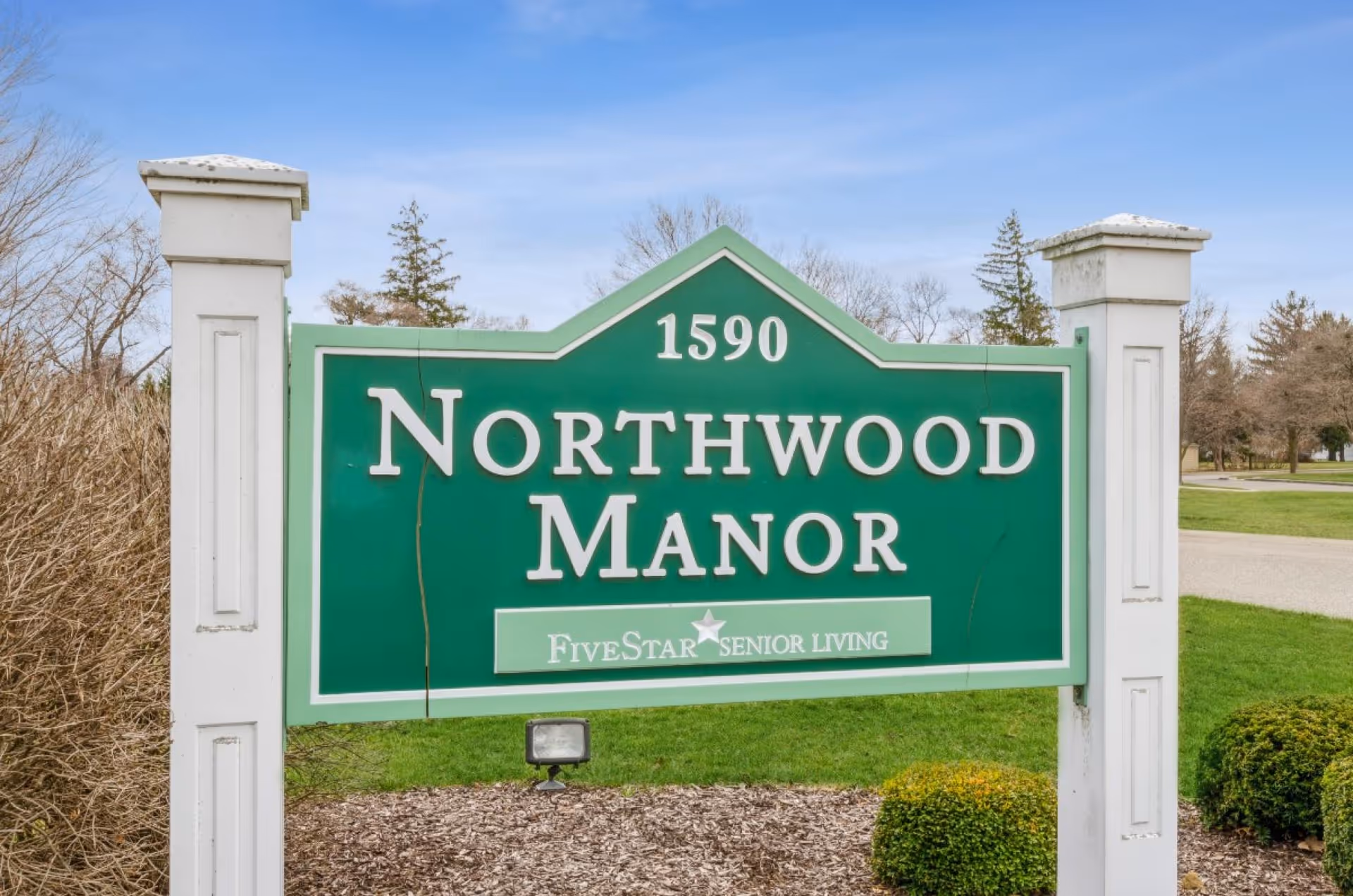 Green entrance sign reading "Northwood Manor" with the address 1590 mounted between white posts on a lawn.