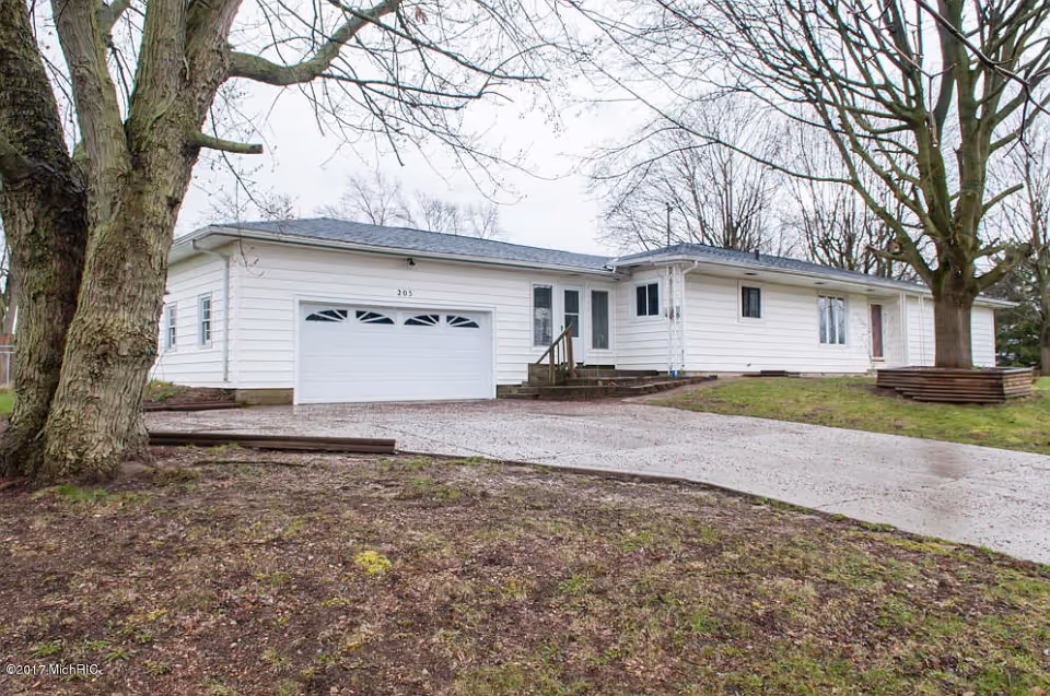 Exterior view of a single-story white house with a two-car garage, a concrete driveway, and leafless trees in the front yard on a cloudy day.