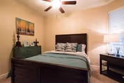 Cozy bedroom with a wooden bed dressed in teal bedding, nightstand with lamp, ceiling fan, and framed wall art.