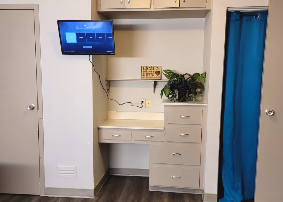 Small interior alcove with a built-in desk and drawers, a wall-mounted TV, a potted plant, and a blue curtain over a doorway.