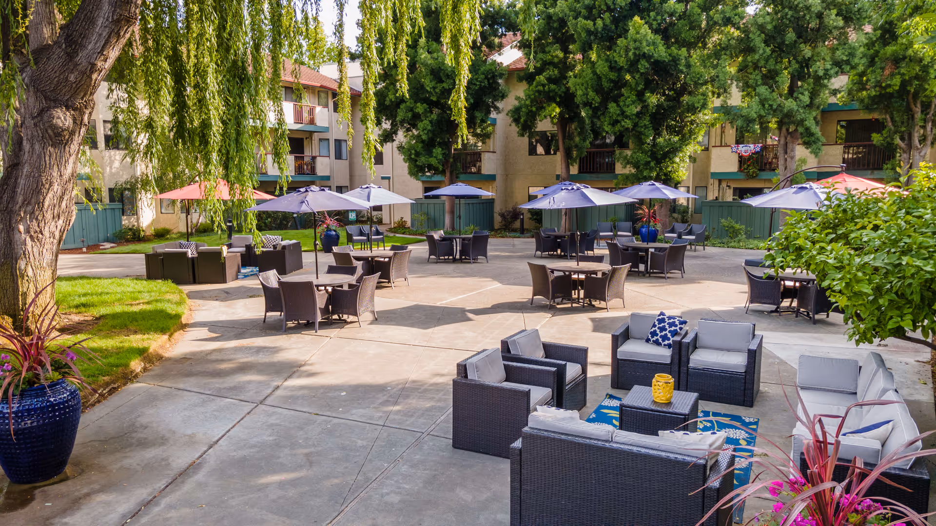 Outdoor patio area at Cogir of Fremont with multiple seating arrangements including wicker chairs and sofas with cushions, round tables with umbrellas, surrounded by trees and apartment buildings in the background.