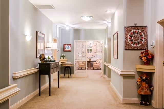 A well-lit hallway in a senior living facility with light-colored walls and carpeted floor. The hallway features decorative wall moldings, framed artwork, a small black console table with a lamp and decorative items, and a small table with a plant. At the end of the hallway, double glass doors lead to a room with two upholstered chairs and a table. On the right side near the foreground, there is a small shelf with a vase of flowers and a decorative doll dressed in autumn attire. The overall atmosphere is warm and inviting.