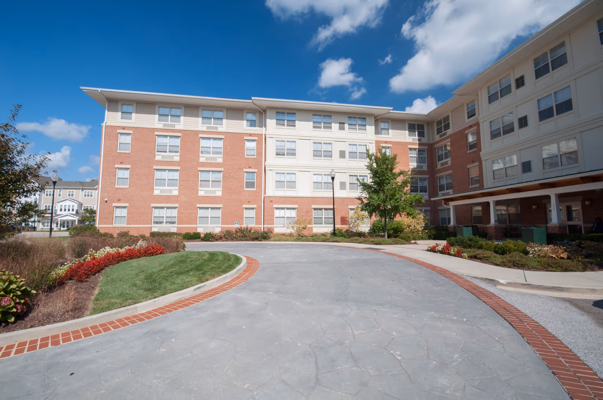 Exterior view of Ednor Apartments II, a multi-story residential building with red brick and beige siding. The building is surrounded by landscaped greenery, including bushes, flowers, and a tree. A curved driveway with a brick border leads up to the entrance under a covered porch. The sky is blue with some clouds.