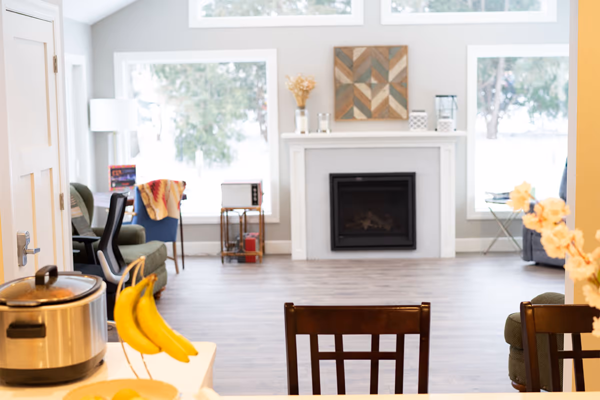 View from a kitchen area looking into a living room with a fireplace centered between two large windows. The room has wooden flooring, a green armchair, a blue chair with a colorful blanket, and a small table with a microwave. Bananas hang on a stand in the foreground on the kitchen counter.