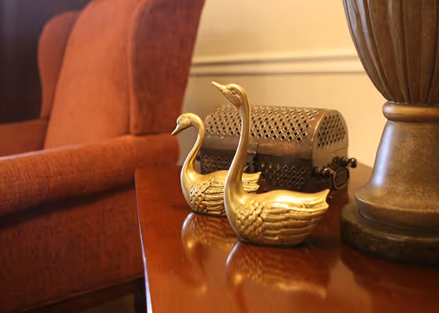 Two brass swan figurines sit on a polished wooden side table next to a lamp with an upholstered chair in the background.