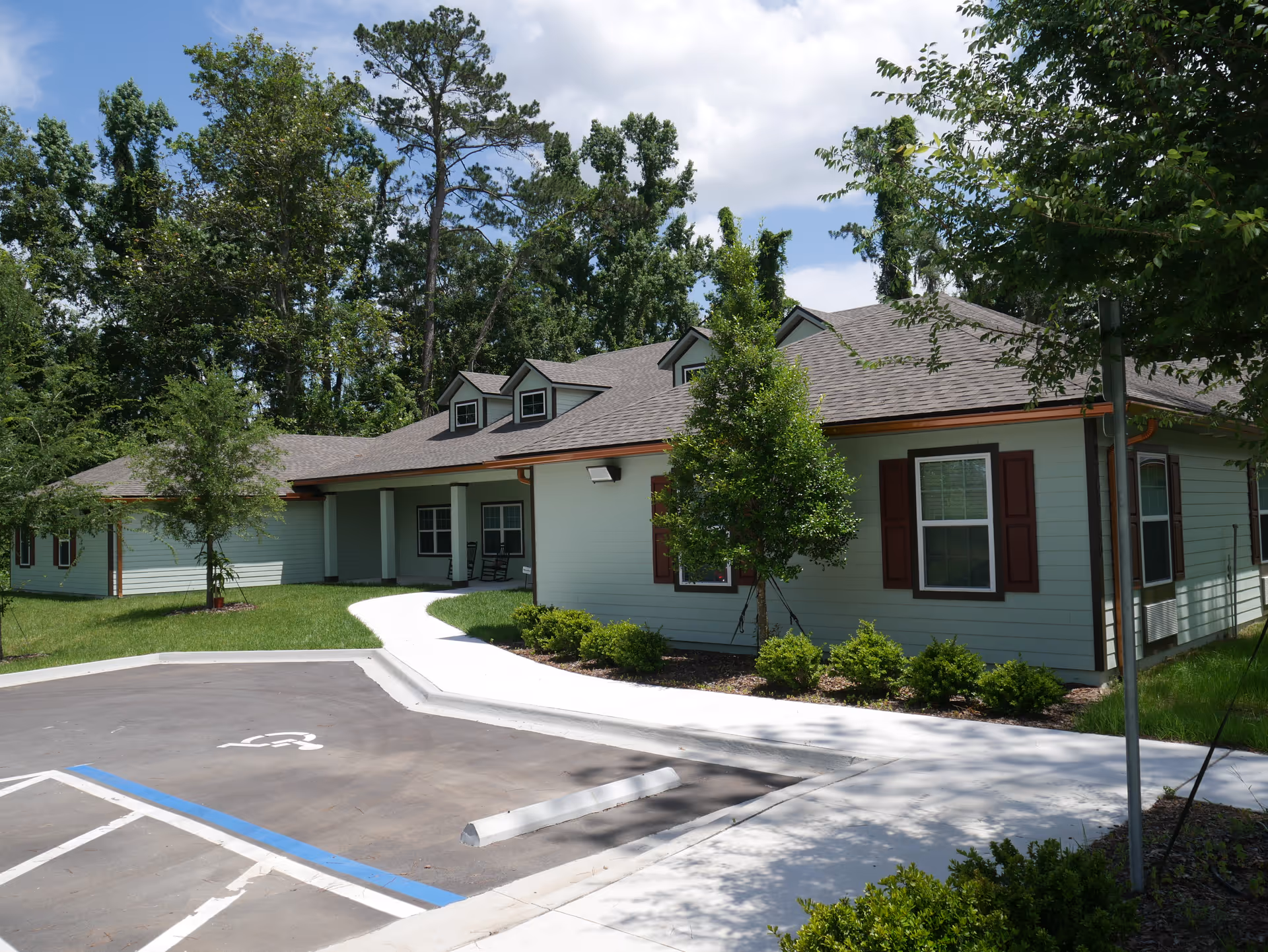Single-story light green senior living building with a covered entrance, walkway, landscaping, and a nearby parking area with a handicapped space.