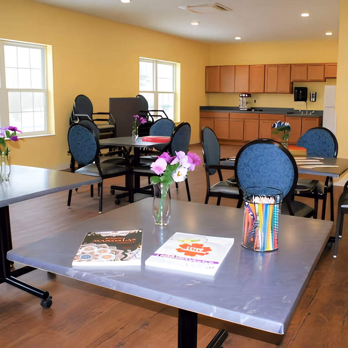 A bright room with several tables and blue cushioned chairs arranged around them. On the tables are vases with purple flowers, books, and a container holding colored pencils. The room has large windows letting in natural light and a kitchen area with wooden cabinets and a refrigerator in the background.