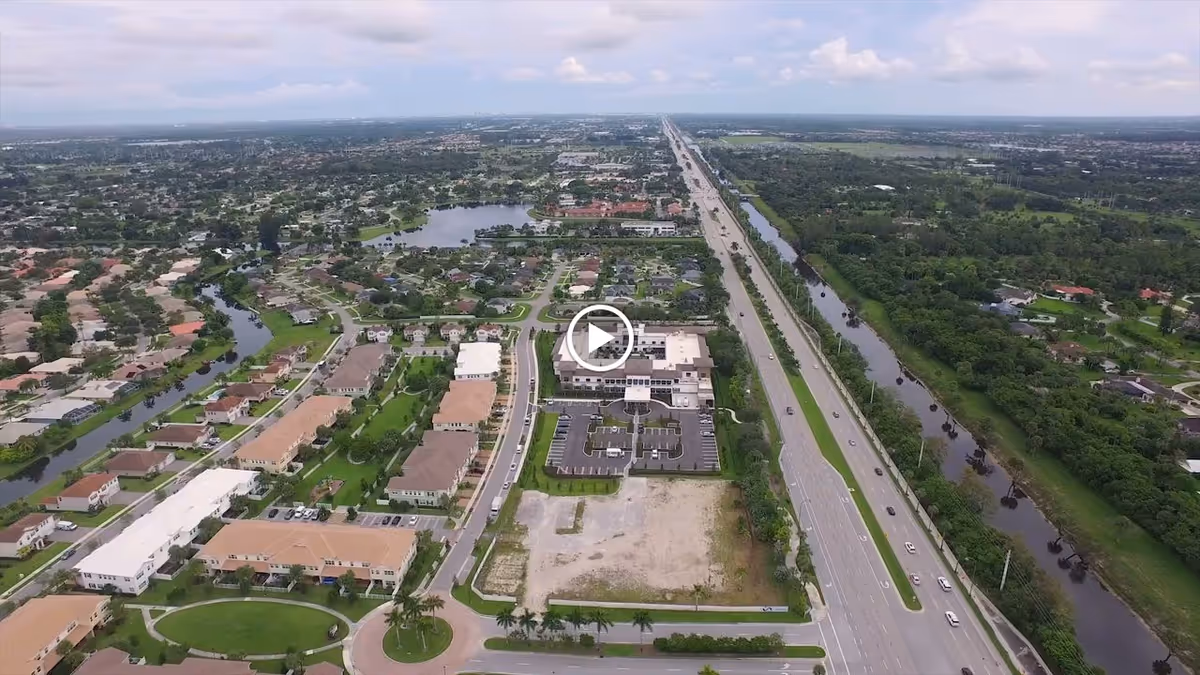 Aerial view of a suburban area featuring residential houses, a large building with a parking lot, a major highway running parallel to a canal, and surrounding greenery under a cloudy sky.