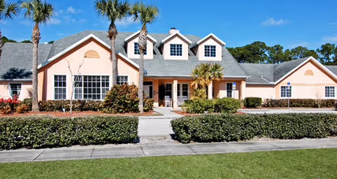 Exterior front view of a single-story senior living facility building with peach-colored walls, multiple windows, a gray shingled roof, and palm trees in front. There is a sidewalk leading to the entrance and well-maintained landscaping with bushes and grass.