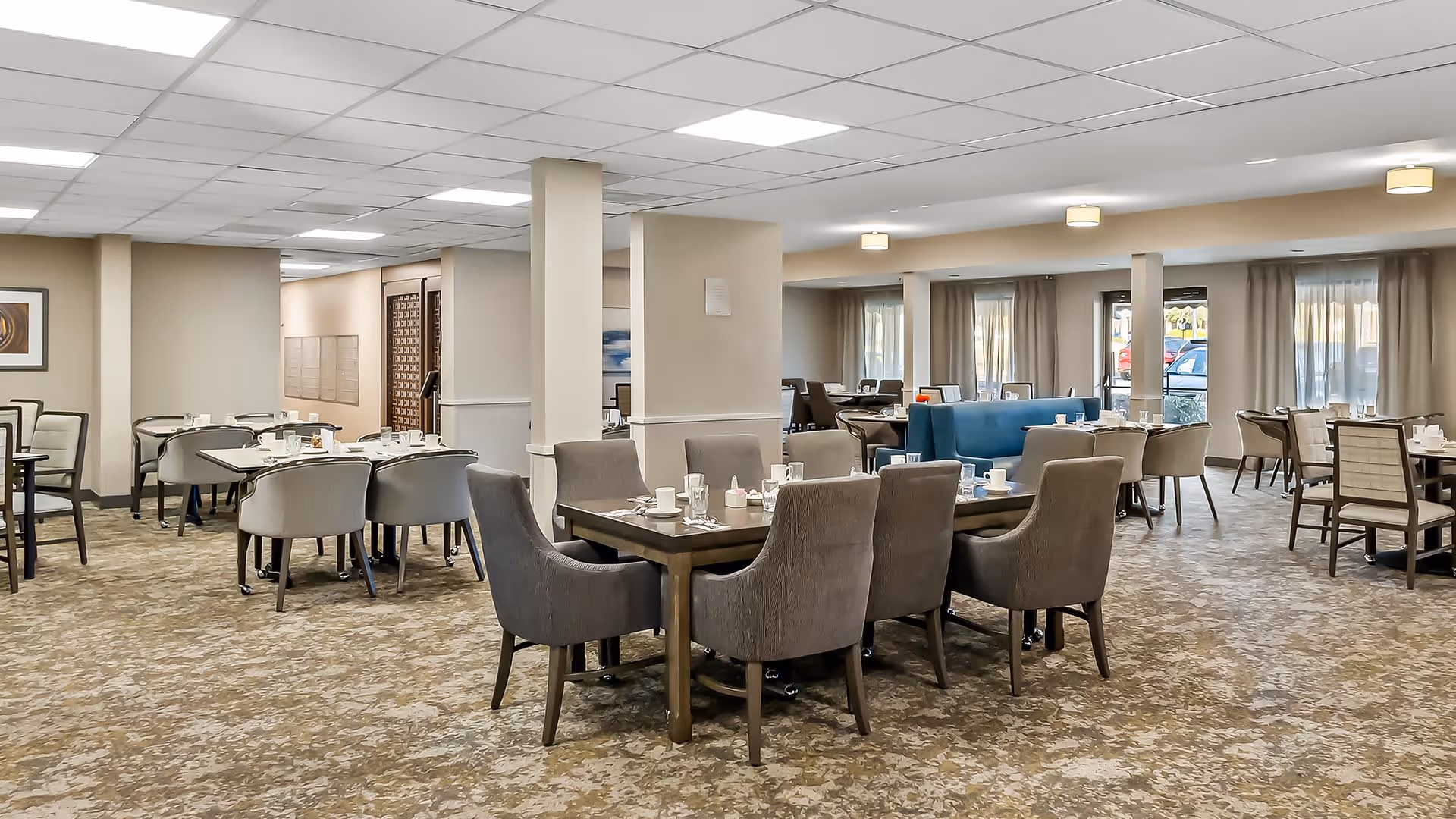 A spacious dining room in a senior living facility with multiple tables and chairs arranged neatly. The tables are set with cups, glasses, and napkins. The room has beige walls, carpeted floors, and large windows with curtains allowing natural light to enter. Ceiling lights provide additional illumination.