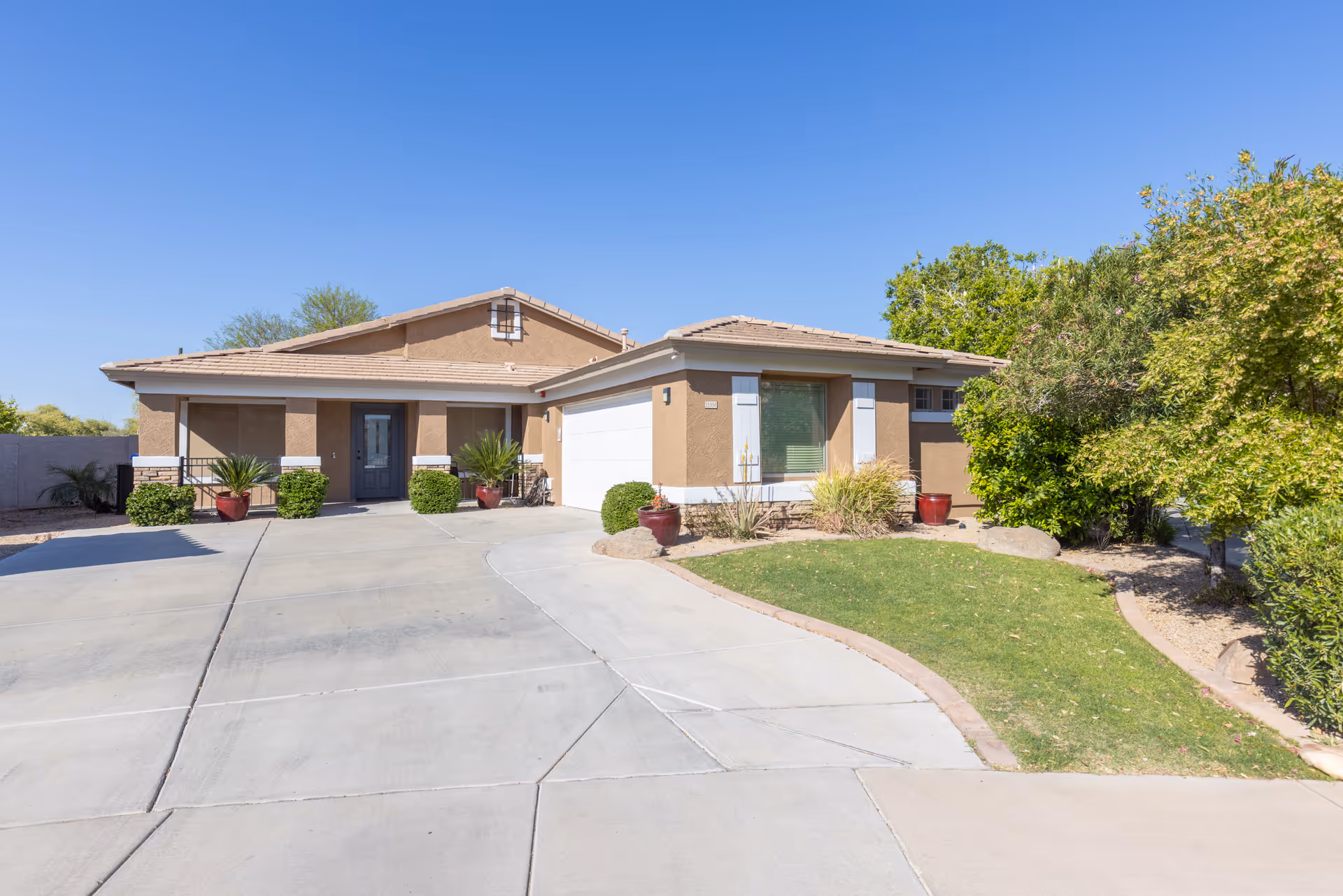 Single-story stucco house with attached garage, driveway, front lawn and potted plants under a clear blue sky.