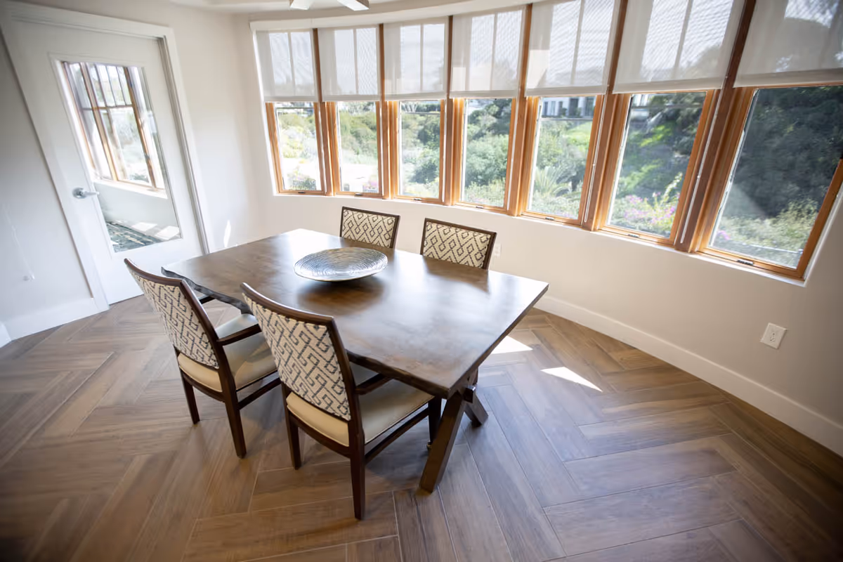Sunlit dining area with a wooden table and four patterned chairs beside a row of large windows overlooking greenery.